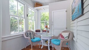Een zonnige kamer met witte muren, een houten vloer, rieten stoelen met blauwe kussens, een klein rond tafeltje en een deur.