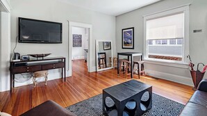 A modern living room with a flat-screen TV, wooden flooring, a dining area with stools, and a large window with blinds. - Old City Charm l Historic l Walk to St George (St. Augustine)