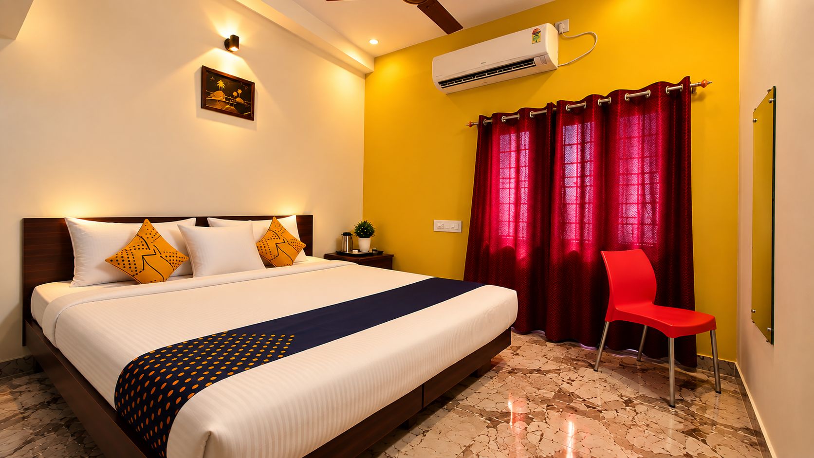 A hotel room with a bed, a nightstand, a red chair, a wall-mounted air conditioner, and a window with red curtains.