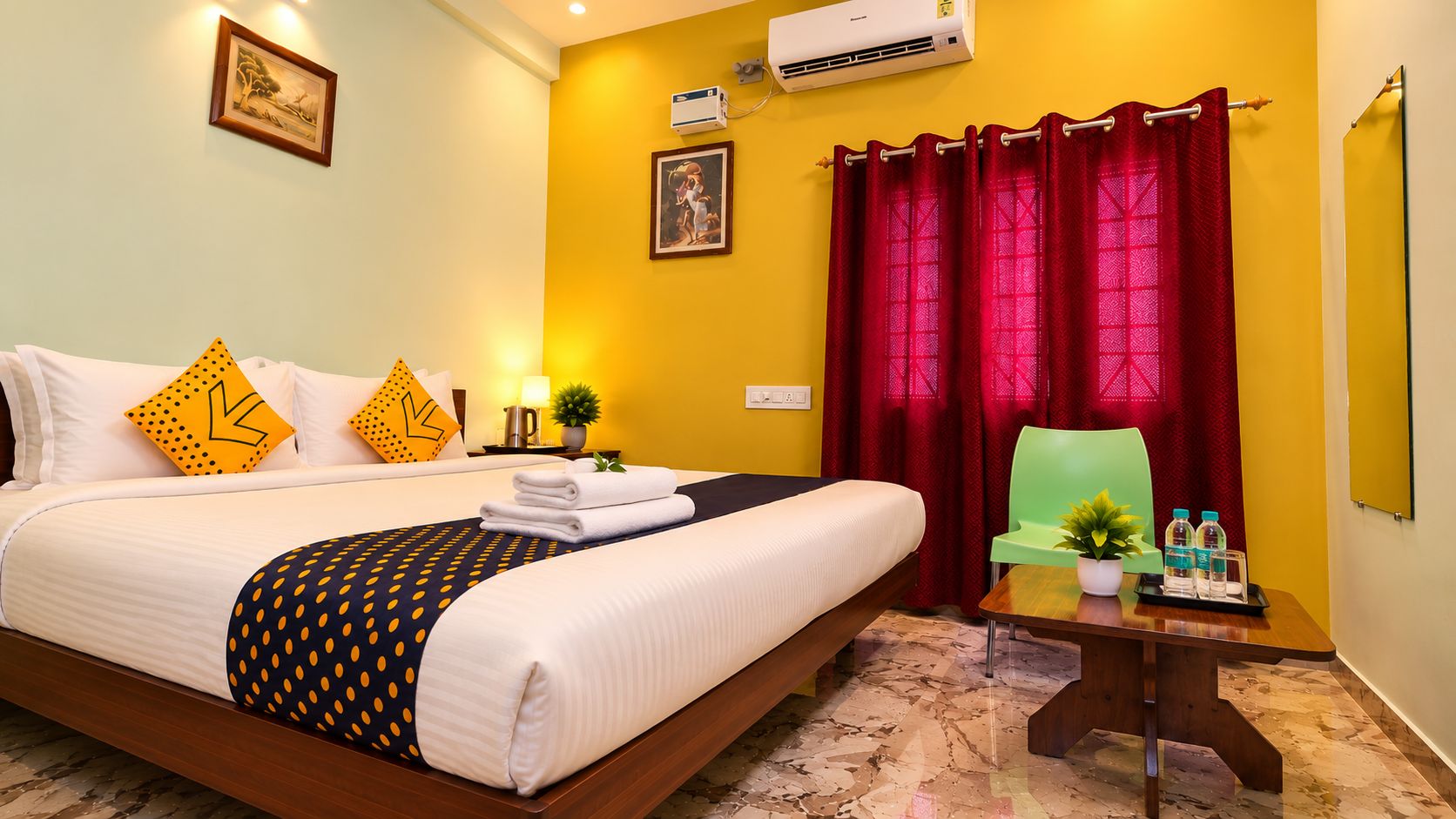 A hotel room with a bed, a desk with a chair, a potted plant, and a red curtain.