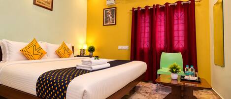 A hotel room with a bed, a desk with a chair, a potted plant, and a red curtain.
