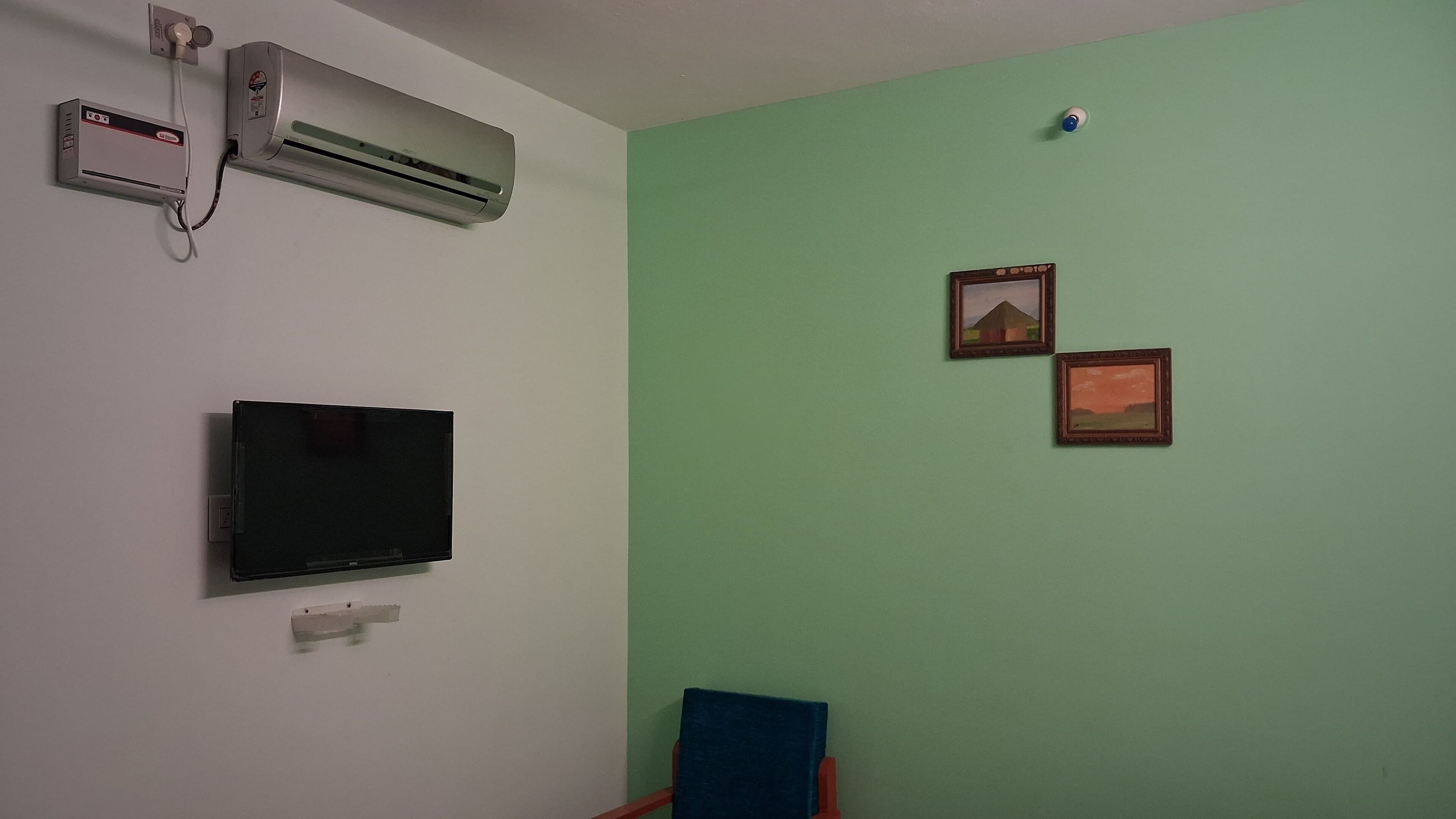 A room with a flat-screen TV mounted on the wall, an air conditioning unit, and two framed pictures on the wall.