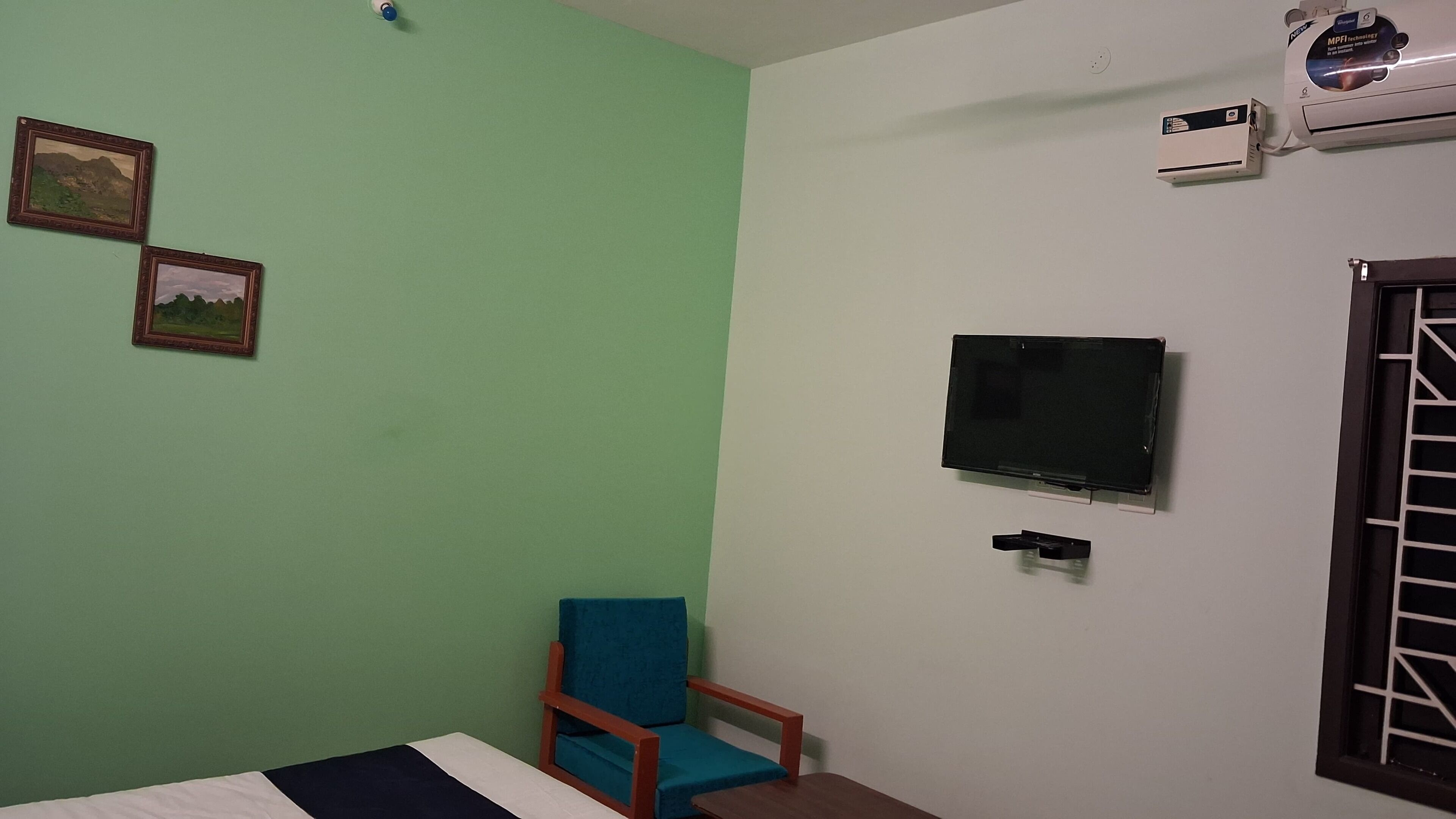 A room with a bed, a blue chair, a TV mounted on the wall, and two air conditioning units.