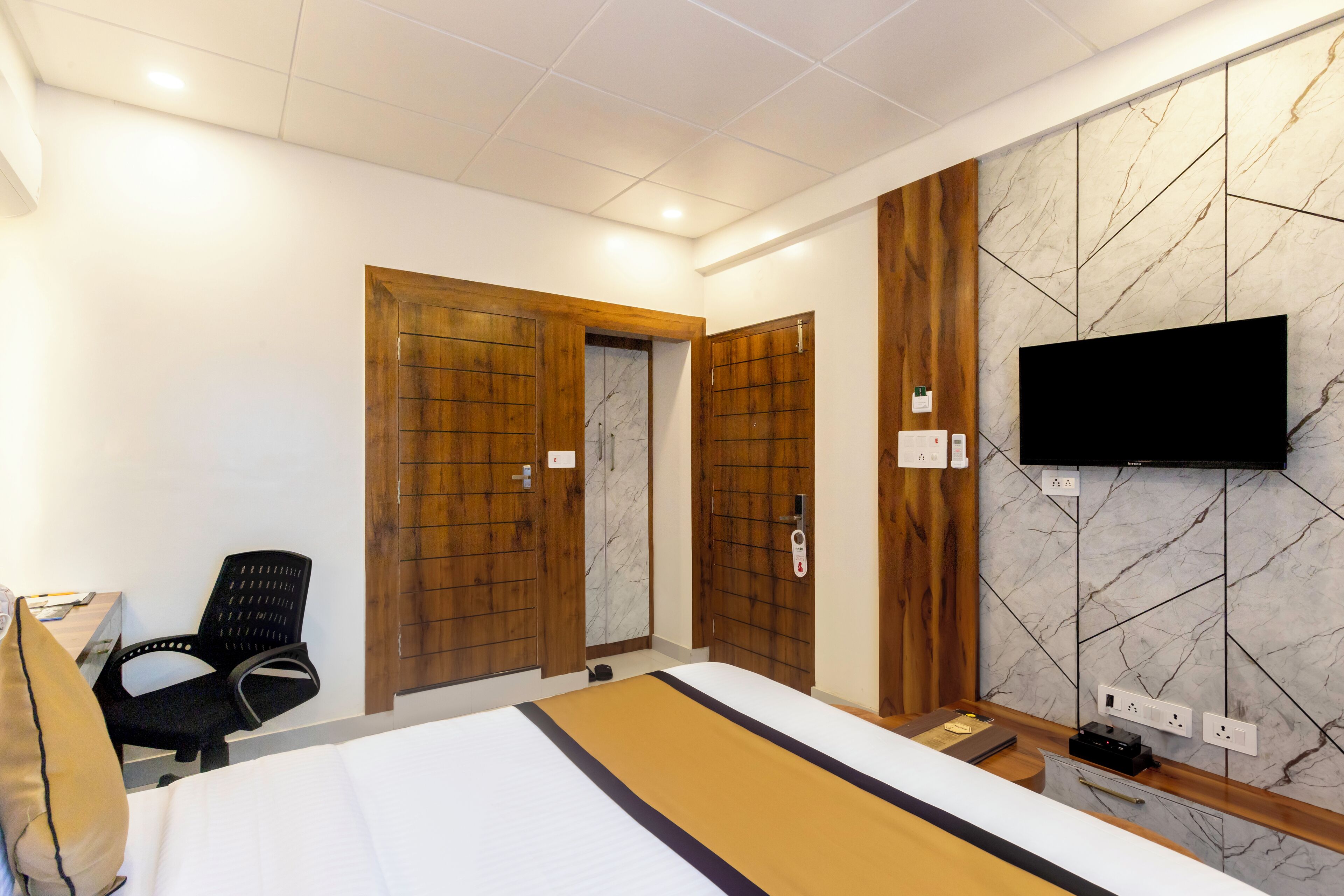 A hotel room with a bed, a desk, a chair, a TV mounted on the wall, and wooden doors.