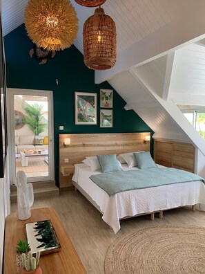 Ein Schlafzimmer mit Holz-Kopfteil, einem Bett mit weißer Bettwäsche, einer gewebten Leuchte und Blick auf einen Balkon mit Pflanze.