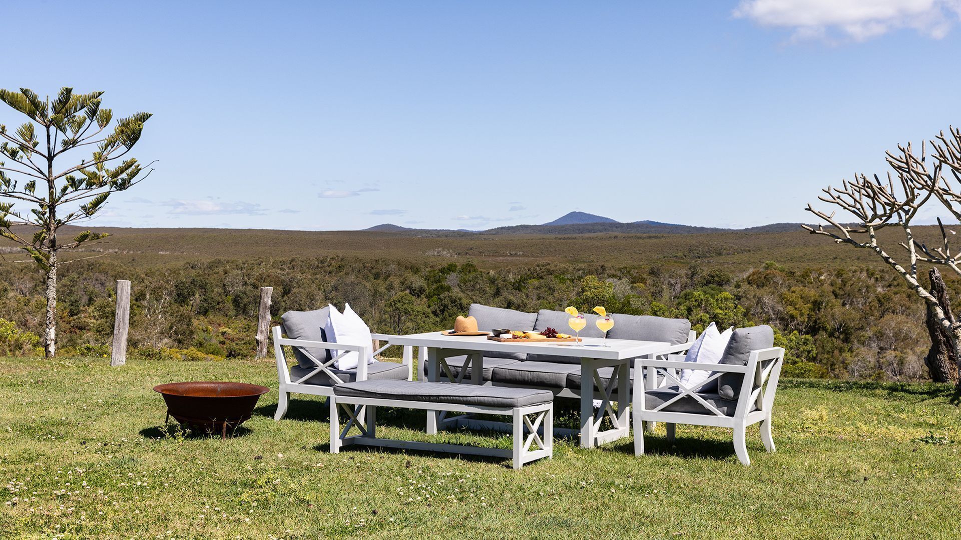 An outdoor seating area with a table and chairs, surrounded by a grassy field and trees, with a view of distant hills.