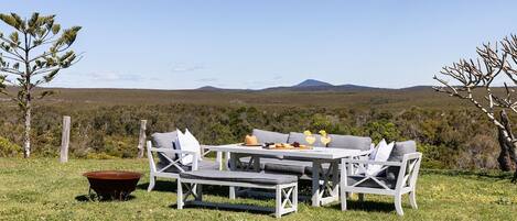 An outdoor seating area with a table and chairs, surrounded by a grassy field and trees, with a view of distant hills.