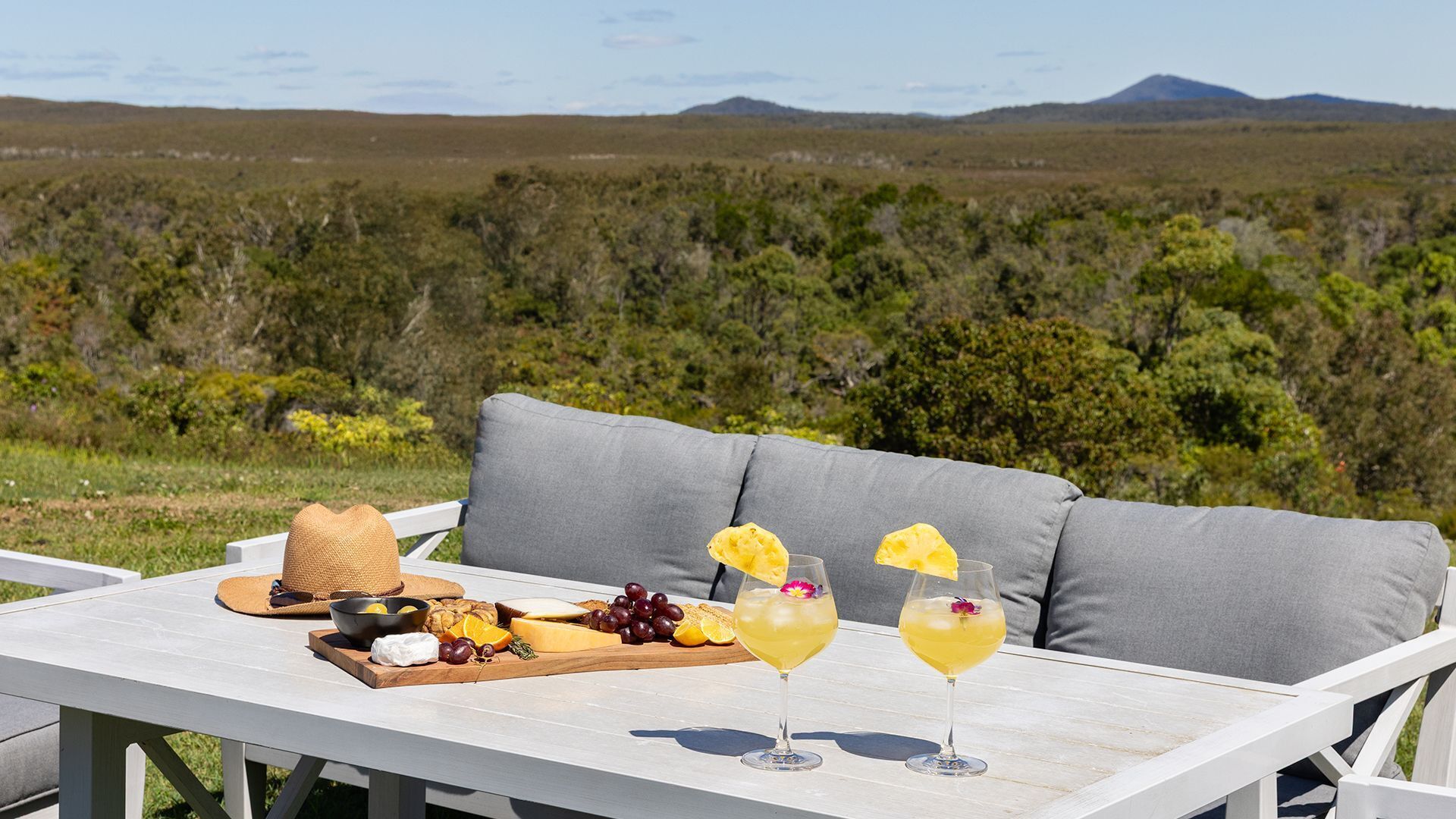 A picnic setup with a table, a white sofa, and a scenic view of a grassy field with trees and mountains in the background.