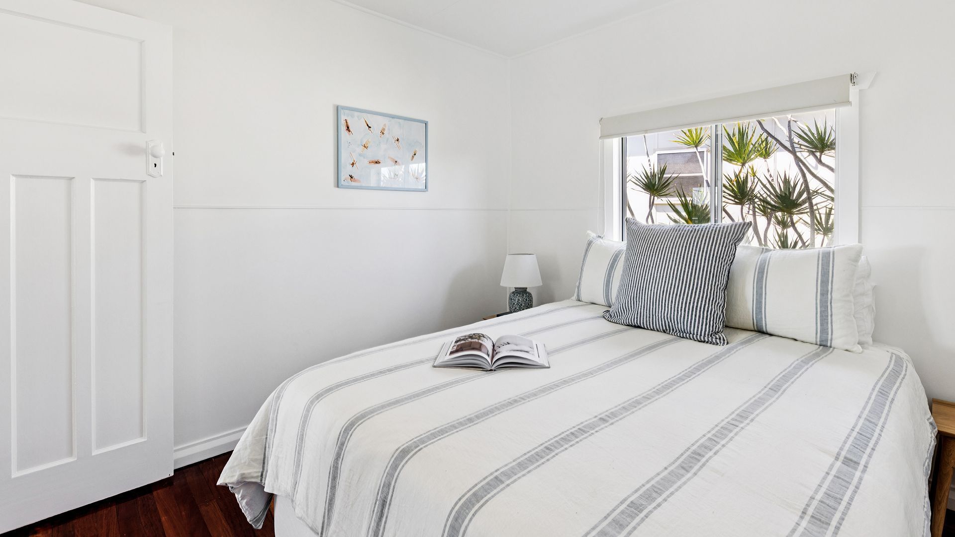 A bedroom with a bed, a window with plants, a door, and a wall-mounted picture.