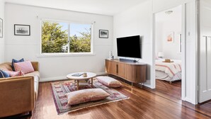 A modern living room with a sofa, a coffee table, a TV, and a view of trees through a large window. - Salt Shack - Classic Angourie Beach Cottage Steps from Spooky Beach - Holiday Yamba (Angourie)