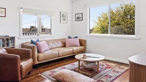 A living room with a brown leather sofa, a round coffee table, a bookshelf, and a large window with a view of trees. - Salt Shack - Classic Angourie Beach Cottage Steps from Spooky Beach - Holiday Yamba (Angourie)