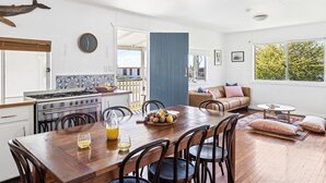 A modern kitchen with a stove, dining table, and chairs. There is a blue door, a sofa, and a window with a view of trees. - Salt Shack - Classic Angourie Beach Cottage Steps from Spooky Beach - Holiday Yamba (Angourie)