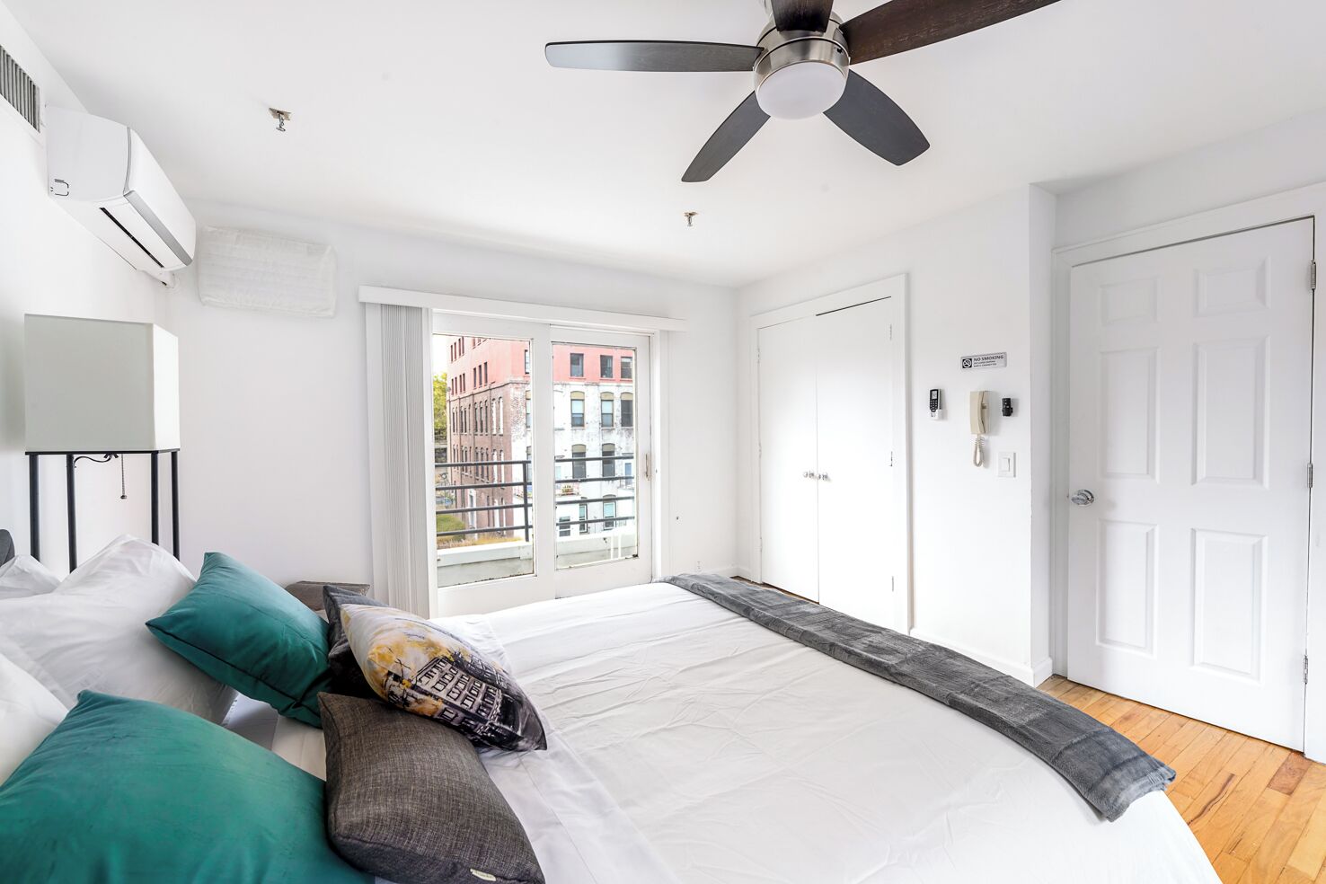 A modern bedroom with a large bed, a ceiling fan, an air conditioning unit, a window with a view of buildings, and a door with a phone and a sign.