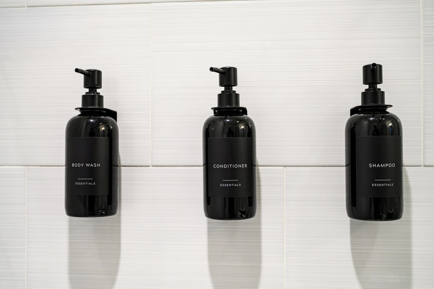 Three black bottles of body wash, conditioner, and shampoo with pump dispensers, labeled 'ESSENTIALS,' against a white tiled background.