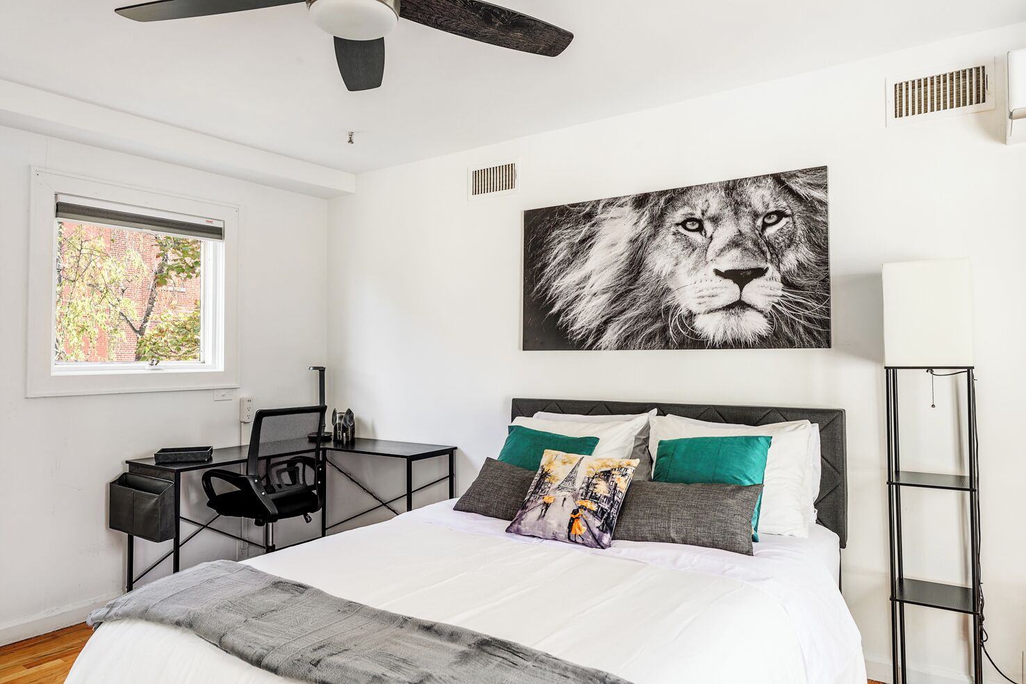 A bedroom with a bed, a desk, a chair, a standing lamp, and a ceiling fan.