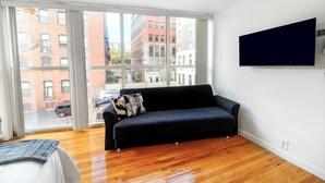 A modern living room with a black sofa, a television mounted on the wall, and a large window offering a view of the city. - Modern 3BR Townhouse with Private Rooftop & Parking Minutes to NYC (Hoboken)