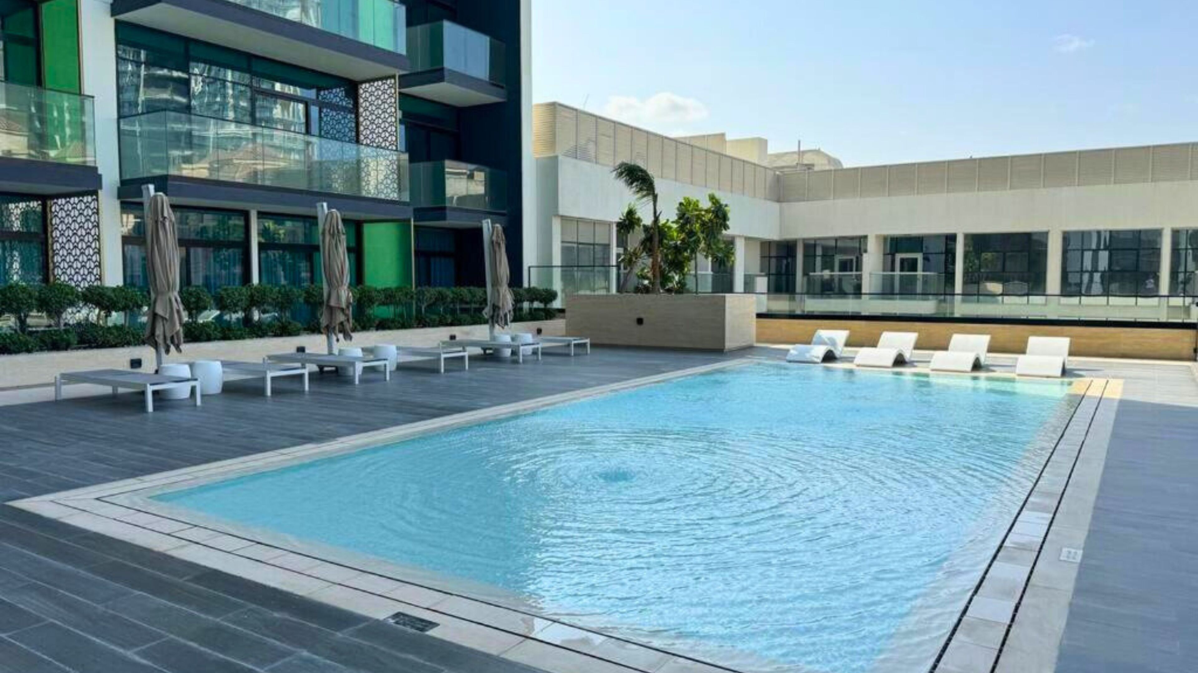 A modern pool area with lounge chairs and umbrellas, surrounded by contemporary buildings.