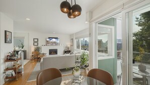 A modern living room with a glass dining table, a sofa, and a fireplace. There is a kitchen area in the background with a TV mounted above the counter. - Artsy Capitol Hill Gem City View Workspace Parking (Seattle)