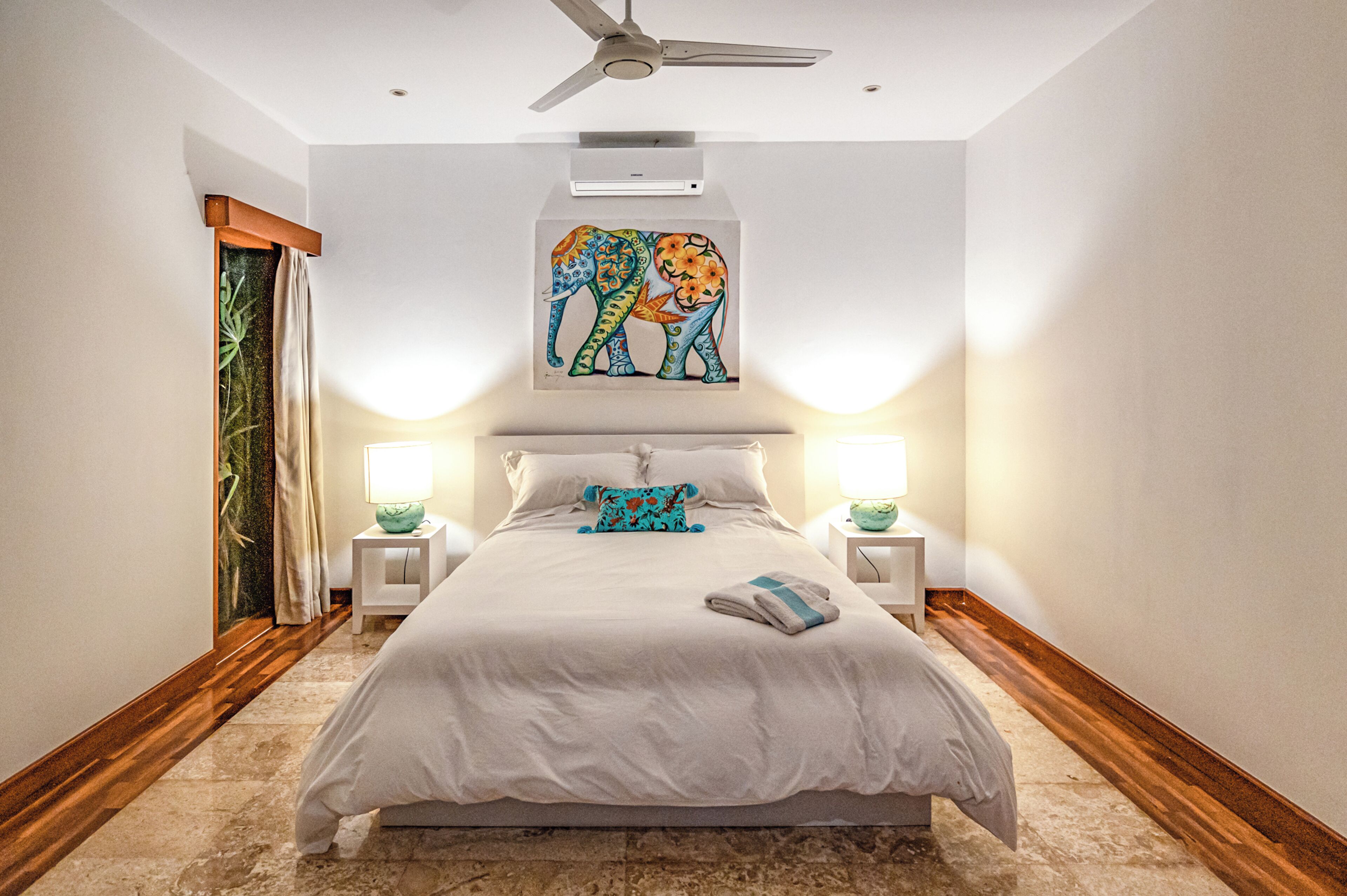 A bedroom with a large bed, two bedside tables, a ceiling fan, and a wall-mounted artwork of elephants.