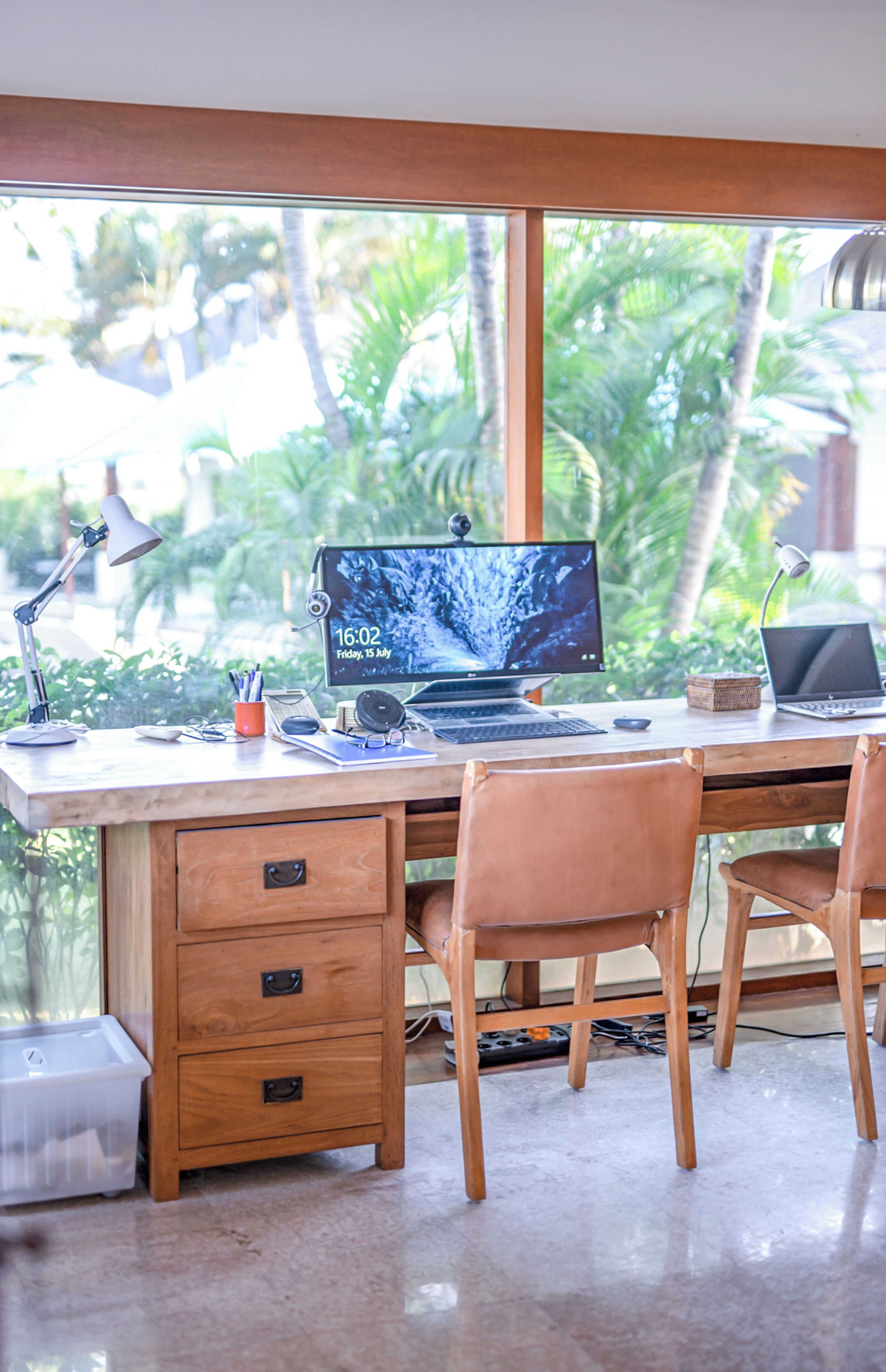 A wooden desk with a laptop, a desktop monitor, and a laptop on a desk with a view of palm trees outside.