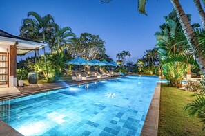 A swimming pool with tiled flooring, lounge chairs, umbrellas, and palm trees. - Villa Mimpi Nyata (Canggu)