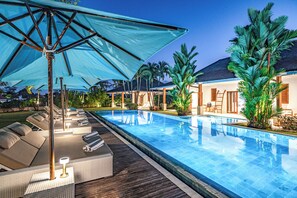 A swimming pool with lounge chairs and umbrellas, surrounded by palm trees and a wooden deck.