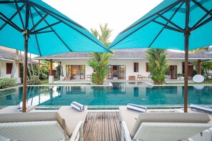 A pool area with lounge chairs and umbrellas, surrounded by a building with a tiled roof and a glass door entrance.