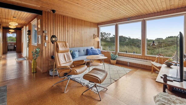 A modern interior with wooden panel walls, a sofa, a chair, a table, and large windows offering a view of the outdoors. - Well-kept 70s holiday home near the North Sea (Hvide Sande)