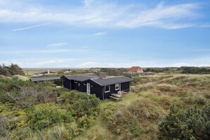 En sort hytte med overdækket terrasse, omgivet af naturlig vegetation og en klar himmel.