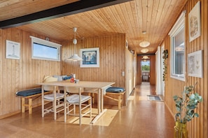 A wooden interior with a dining table, benches, pendant lights, framed artwork, and a wooden floor.