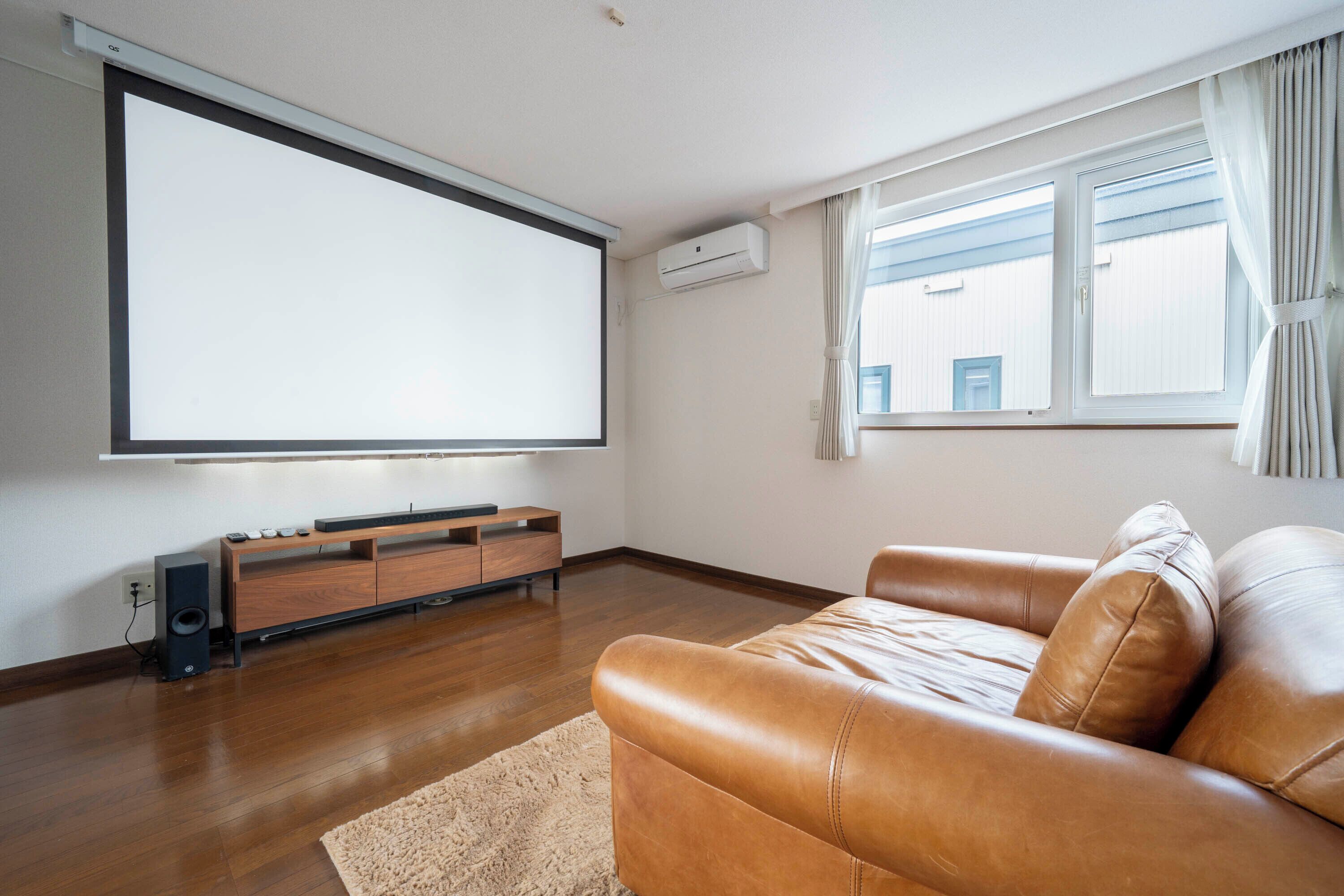 A living room with a large flat-screen TV, wooden flooring, a leather sofa, and a small wooden entertainment unit.