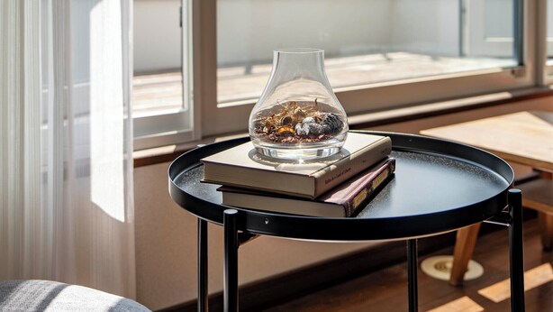 Une pièce dotée d’une grande fenêtre, d’une table ronde noire sur laquelle se trouvent un livre et un vase en verre, et qui offre une vue sur les montagnes et les bâtiments environnants.