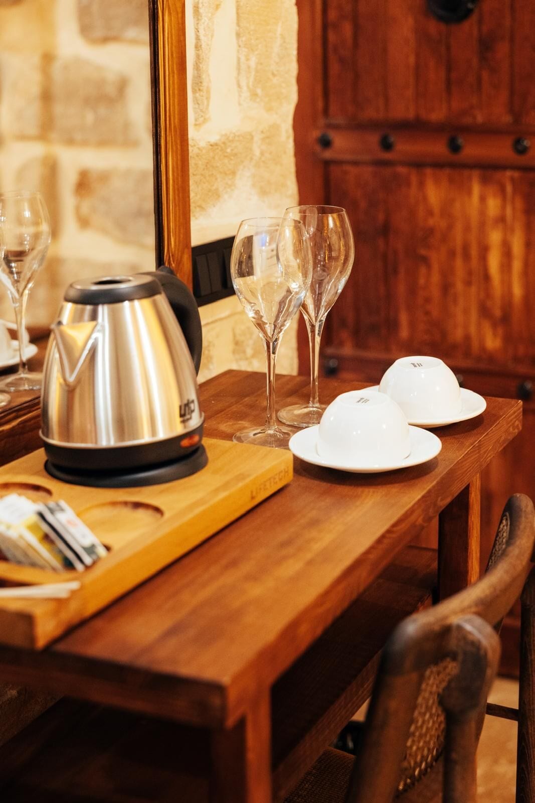 Una mesa de madera con una tetera, copas de vino y platos, frente a una pared de piedra y un armario de madera.