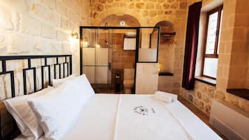 Une chambre d’hôtel avec un mur en pierre, un lit avec du linge de maison blanc, une tête de lit en bois, une fenêtre avec des rideaux et un miroir.