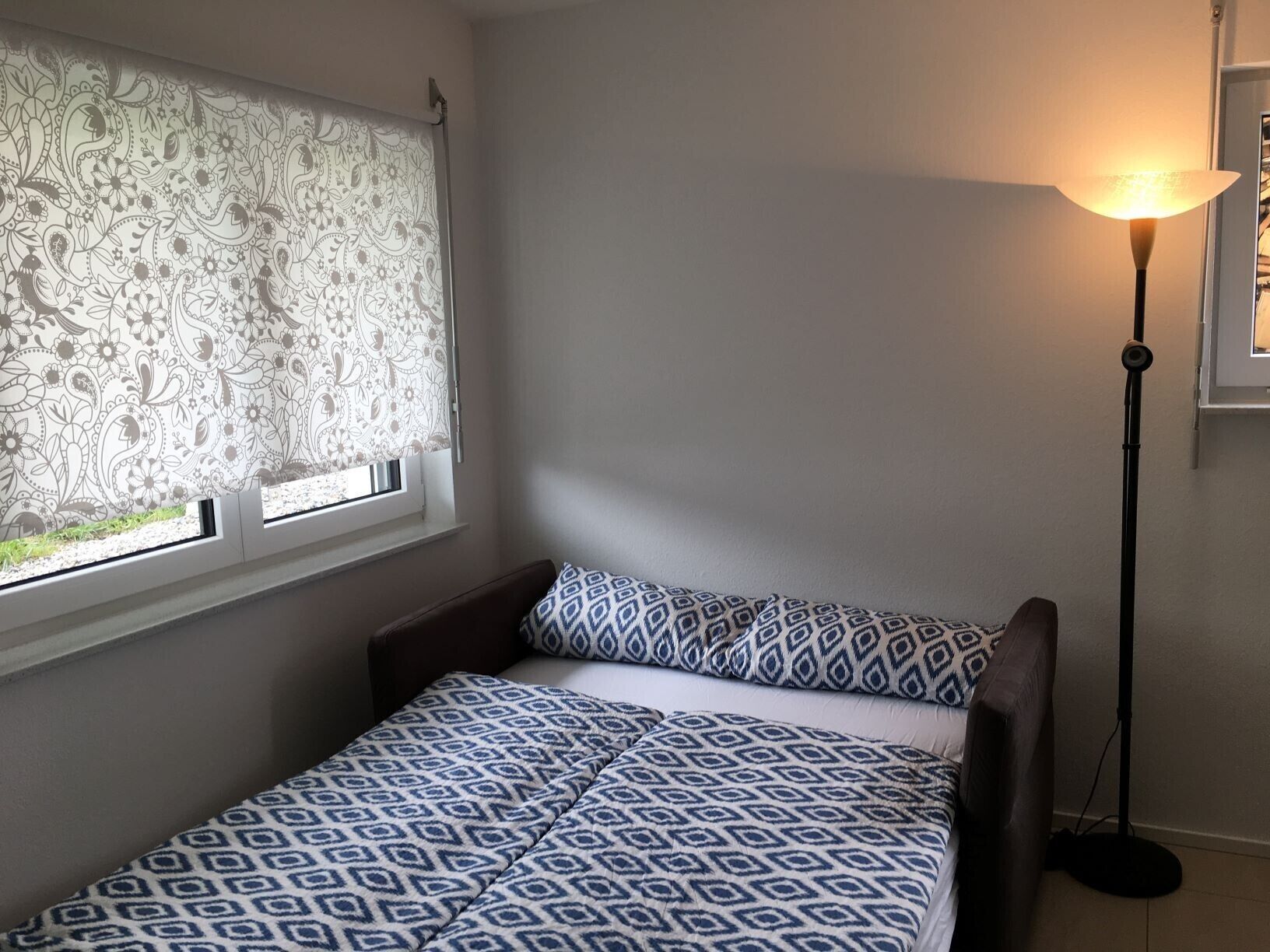 A small bedroom with a window covered by a patterned curtain, a bedside lamp, and a bed with patterned bedding.