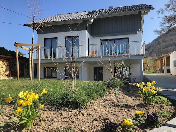 A two-story building with a balcony, a stone wall, and a garden with yellow flowers.