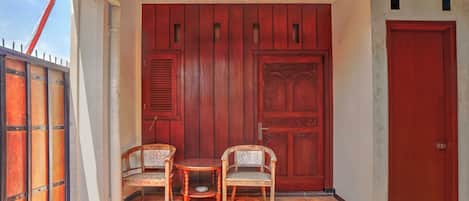 Uma área de estar coberta ao ar livre, com duas cadeiras e uma mesa pequena, uma porta vermelha e um portão de metal.