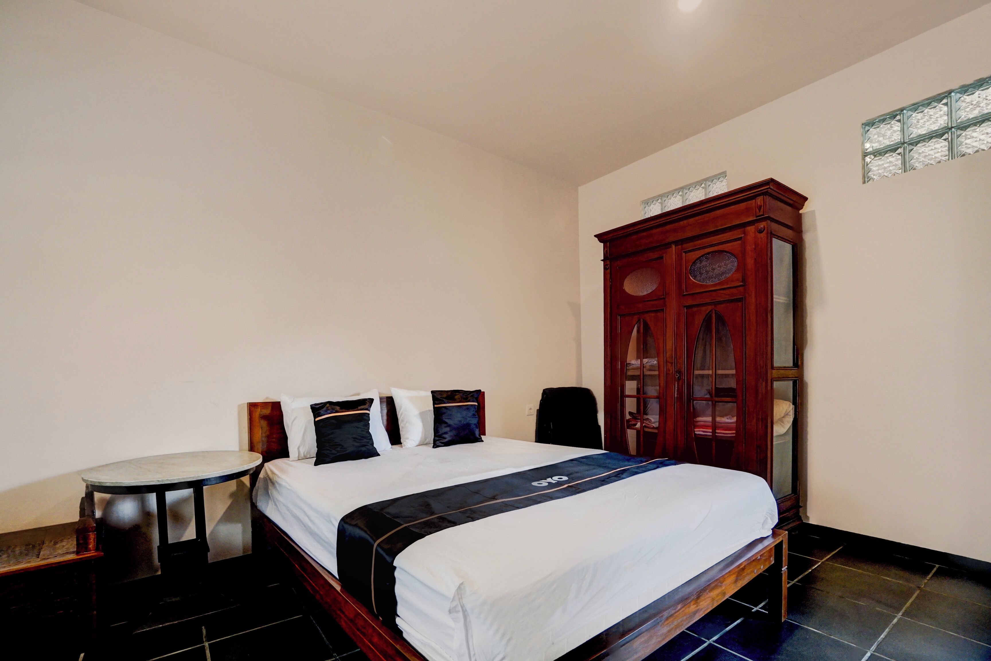 A bedroom with a bed, a wooden wardrobe, a small round table, and a chair.