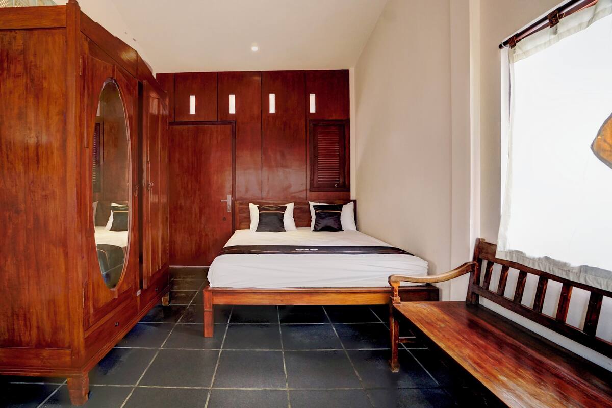 Un dormitorio con un amplio armario de madera, una cama con colchón blanco y almohadas oscuras, un banco de madera y una ventana con cortinas blancas.
