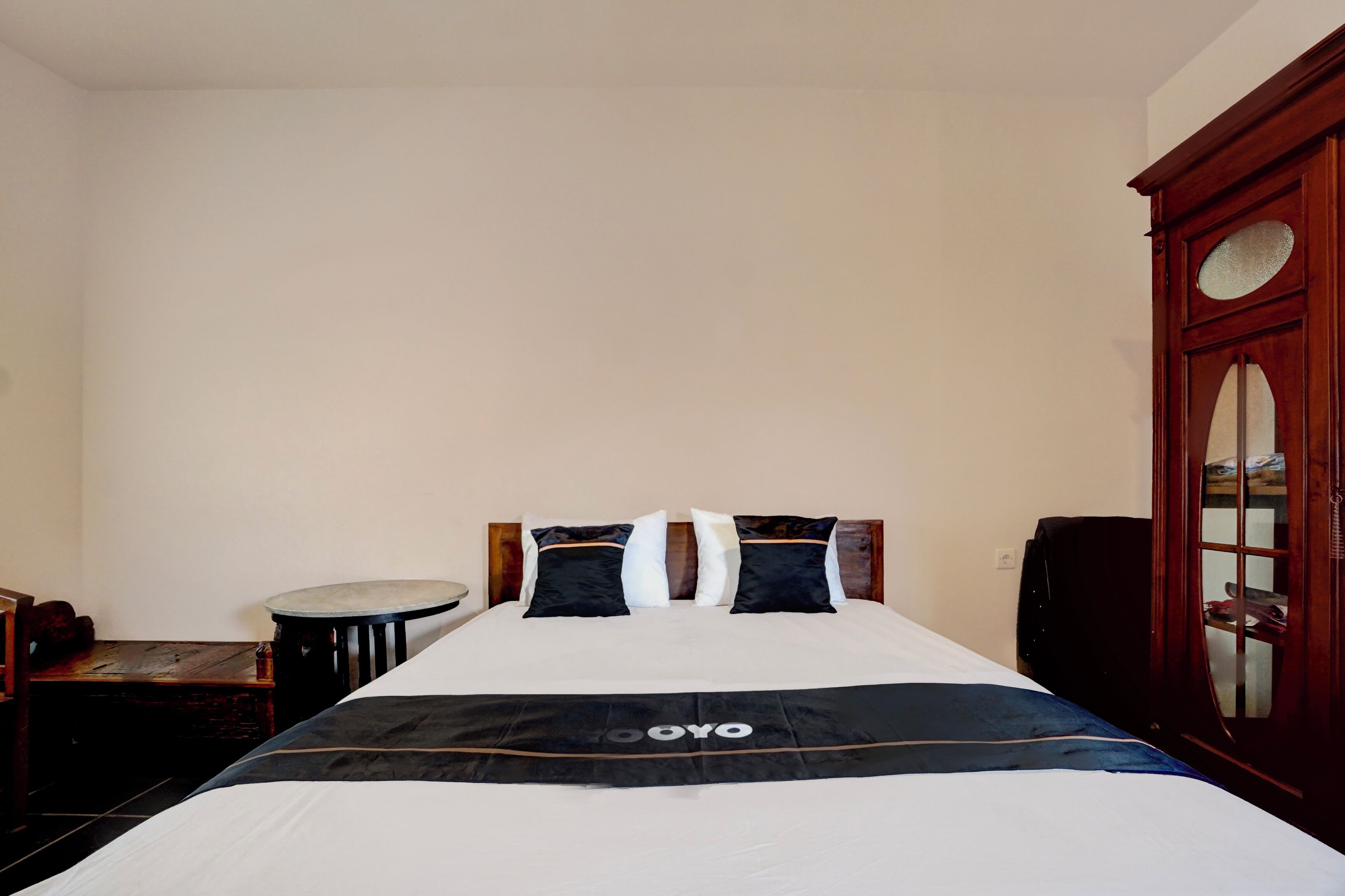 A single bed with a black and white bedspread, a wooden nightstand, a small round table, and a wooden wardrobe with a glass door.