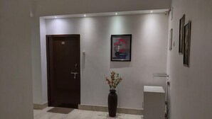 A hallway with a dark wooden door, a framed artwork on the wall, a tall vase with flowers, and a small round table. - The Executive Inn (Guwahati)