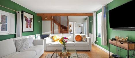 A living room with a white sofa, a coffee table with books, a vase with flowers, and a TV on the wall.