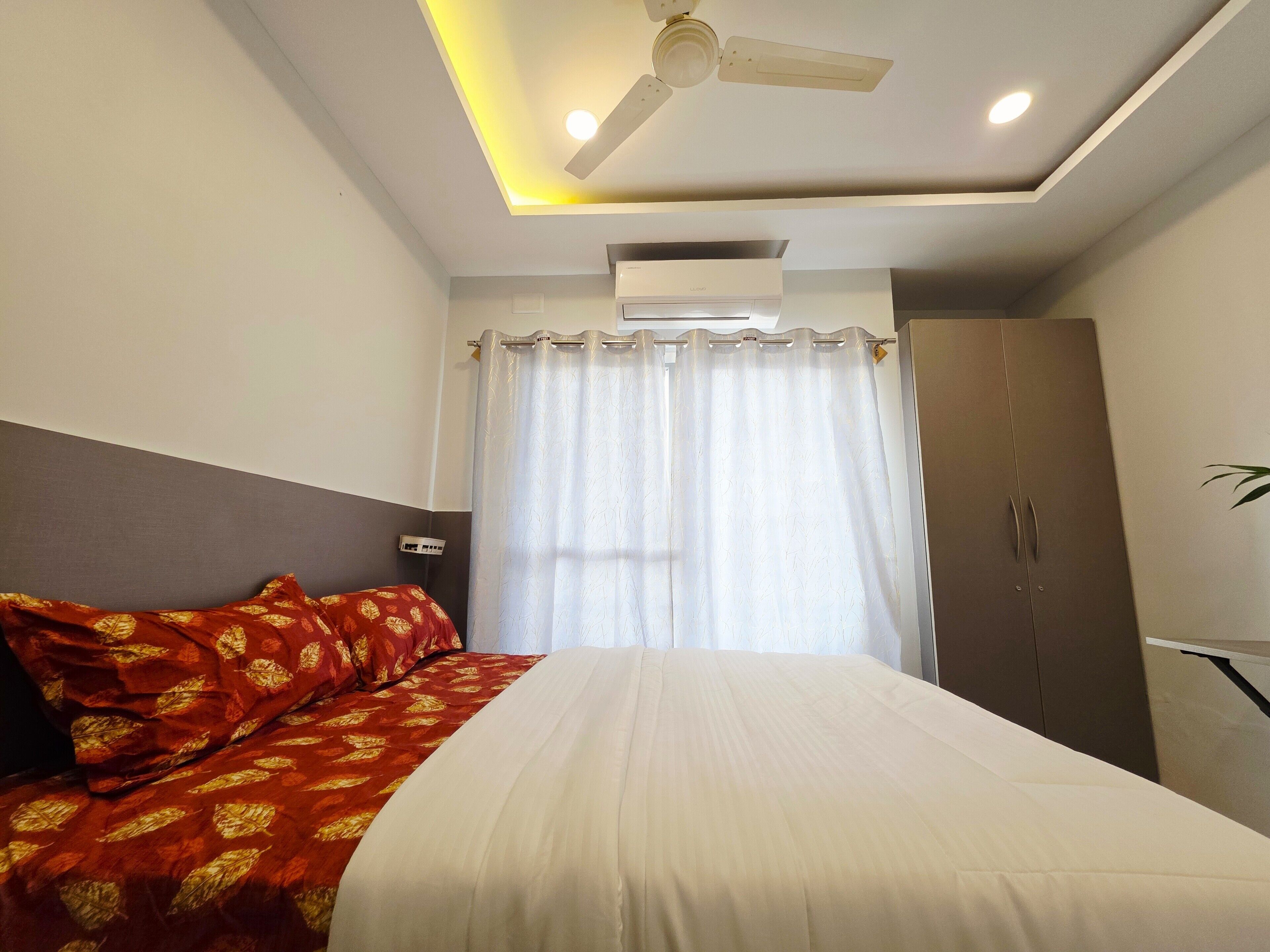 A hotel room with a bed, a ceiling fan, an air conditioning unit, a wardrobe, and a window with curtains.
