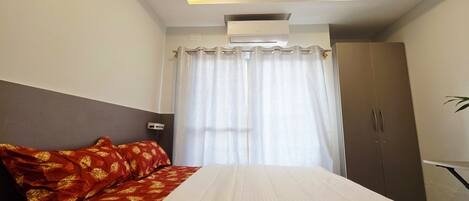 A hotel room with a bed, a ceiling fan, an air conditioning unit, a wardrobe, and a window with curtains.