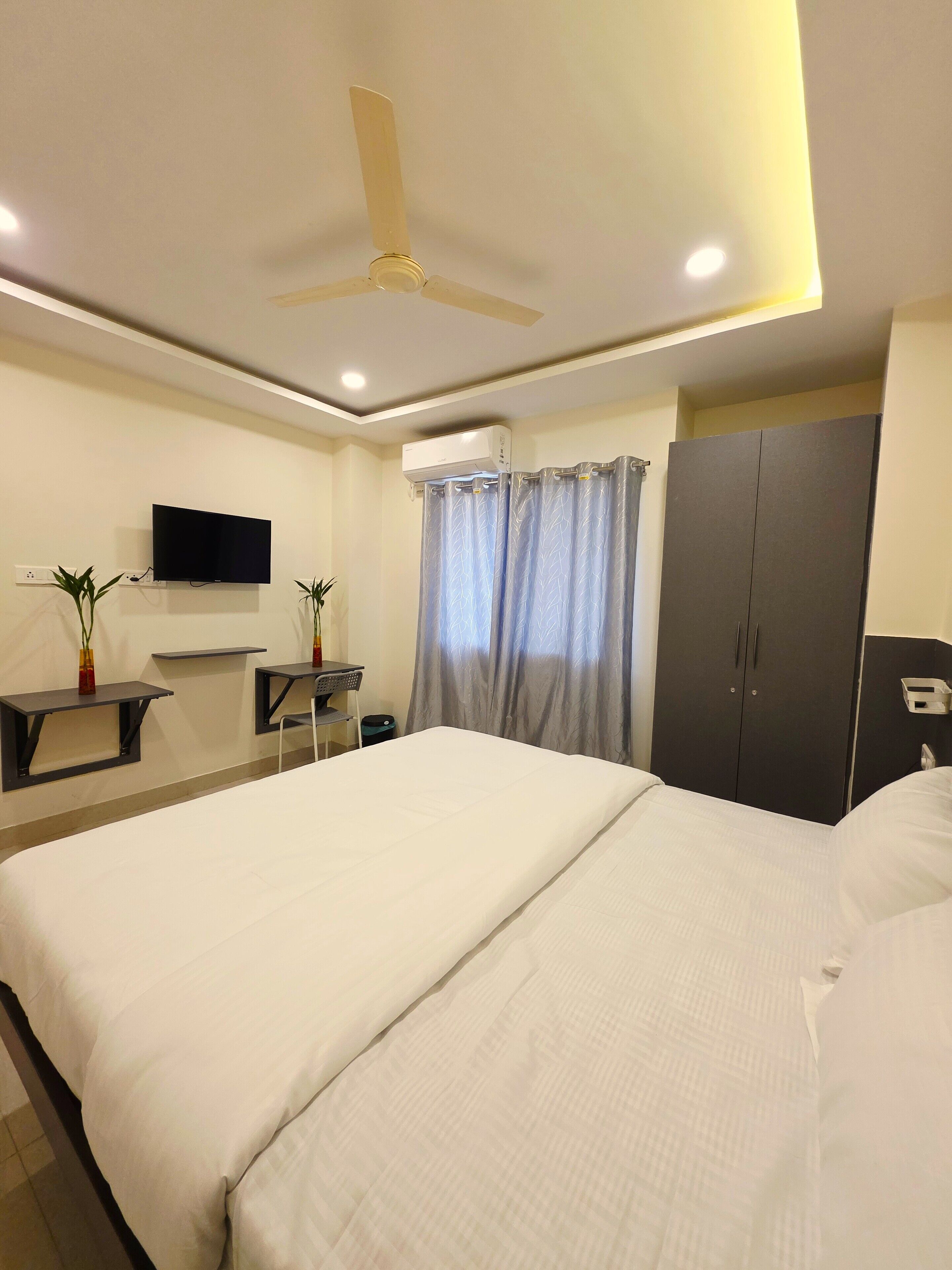 A hotel room with a large bed, a desk with a flat-screen TV, a wardrobe, and a ceiling fan.