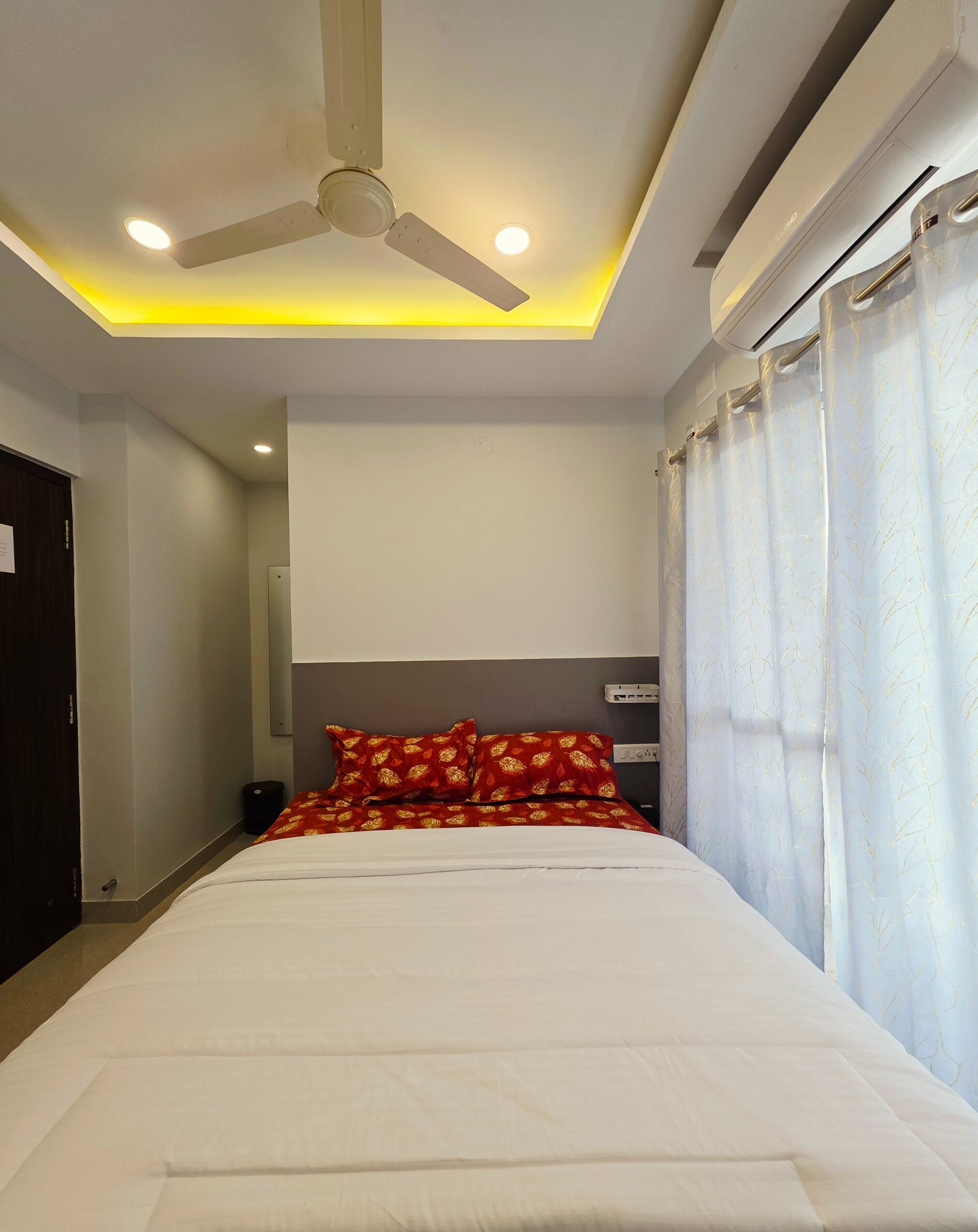 A hotel room with a bed, a ceiling fan, recessed lighting, a window with curtains, and a wall-mounted air conditioning unit.