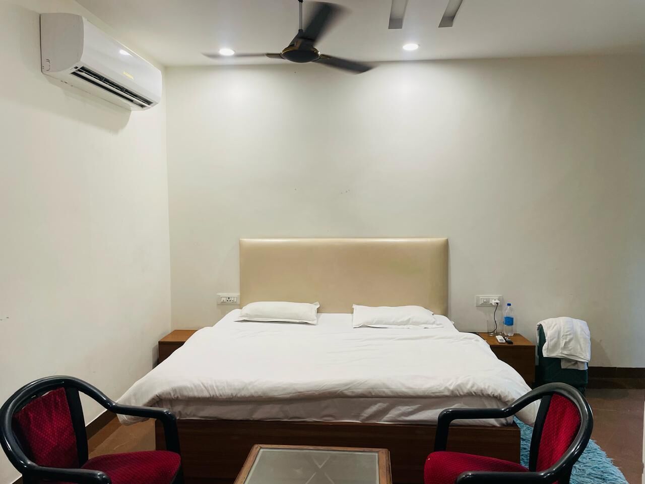 Une chambre d’hôtel comprenant un lit, deux chaises, une table, un climatiseur, un ventilateur de plafond, une table de chevet et une bouteille d’eau.