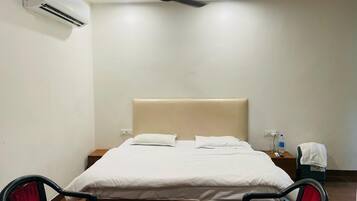 Une chambre d’hôtel comprenant un lit, deux chaises, une table, un climatiseur, un ventilateur de plafond, une table de chevet et une bouteille d’eau.