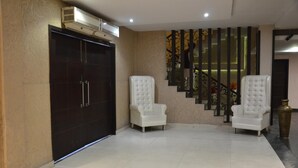 Una habitación amplia con una puerta oscura grande, dos sillones blancos, una escalera con barandillas decorativas y un jarrón alto.