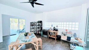 A spacious room with a ceiling fan, a large window with a view of trees, a wooden table with a glass top, and a kitchenette area.